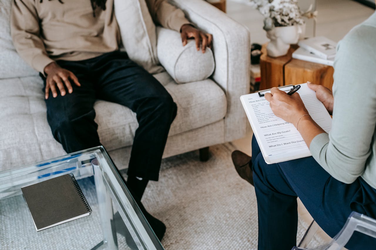 From above crop African American male in casual clothes sitting on comfy couch and answering professional female consultant questions in modern psychotherapy office